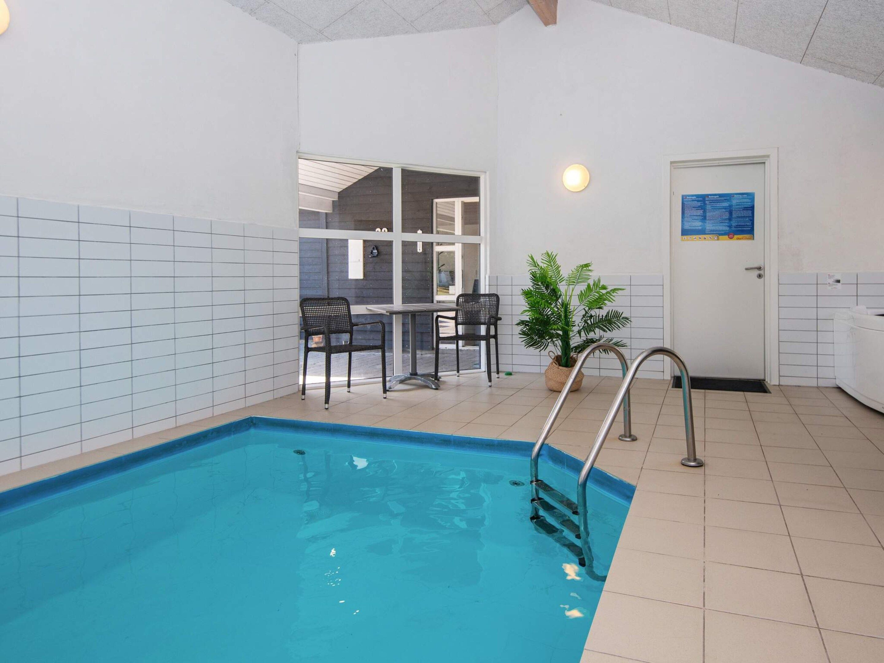 An indoor swimming pool with a metal ladder, two chairs, a potted plant, and a door with a sign.