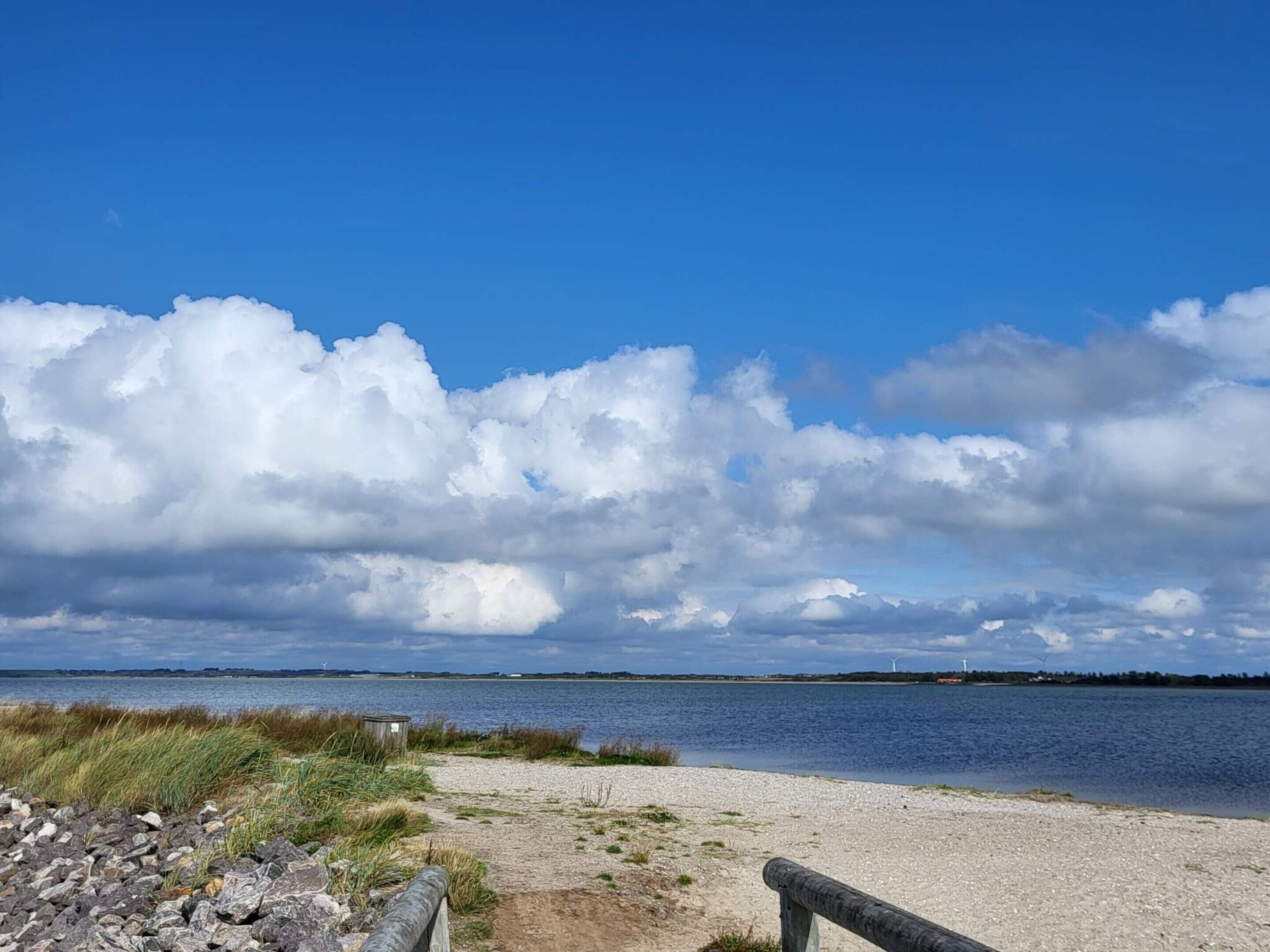 A beach with a gravel path, a wooden fence, and a body of water with distant wind turbines.