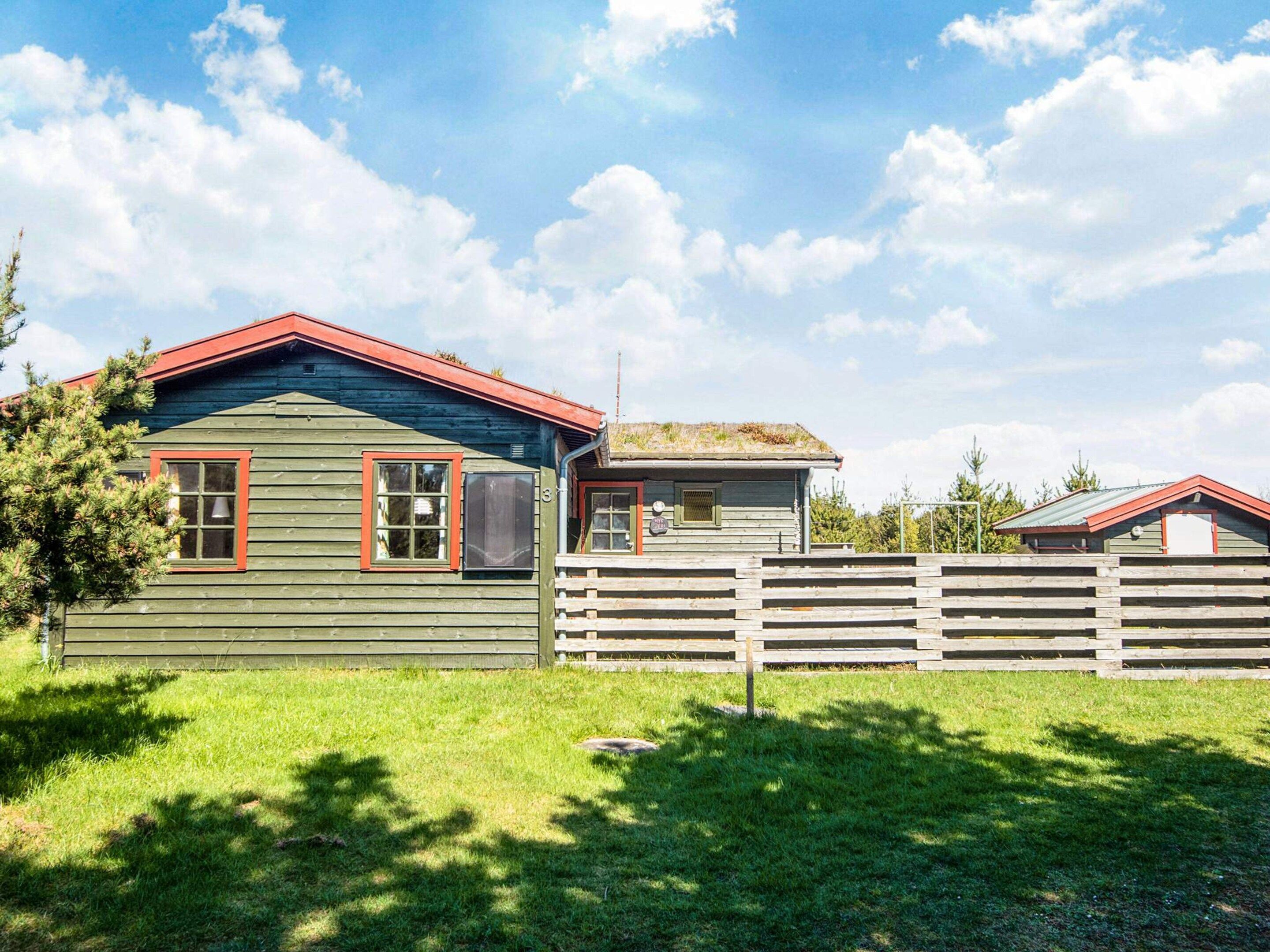 Une cabane rustique à l’extérieur vert et aux bordures rouges, avec une clôture en bois et un ciel dégagé.