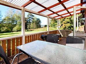 Une terrasse extérieure couverte, équipée d’une table et de chaises, d’un barbecue, et offrant une vue sur un espace verdoyant avec des arbres.