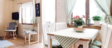 A cozy room with a wooden floor, a dining table with a checkered tablecloth, white chairs with cushions, a television mounted on the wall, and a window with curtains.