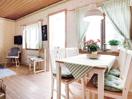 A cozy room with a wooden floor, a dining table with a checkered tablecloth, white chairs with cushions, a television mounted on the wall, and a window with curtains.