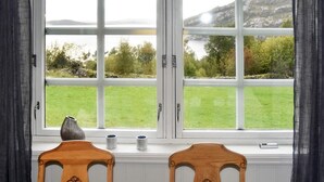 Una habitación con un ventanal grande, dos sillas de madera, un florero y dos tazas sobre el alféizar.