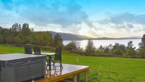 Un jacuzzi en una terraza de madera con una mesa y sillas, con vista a un lago y colinas.