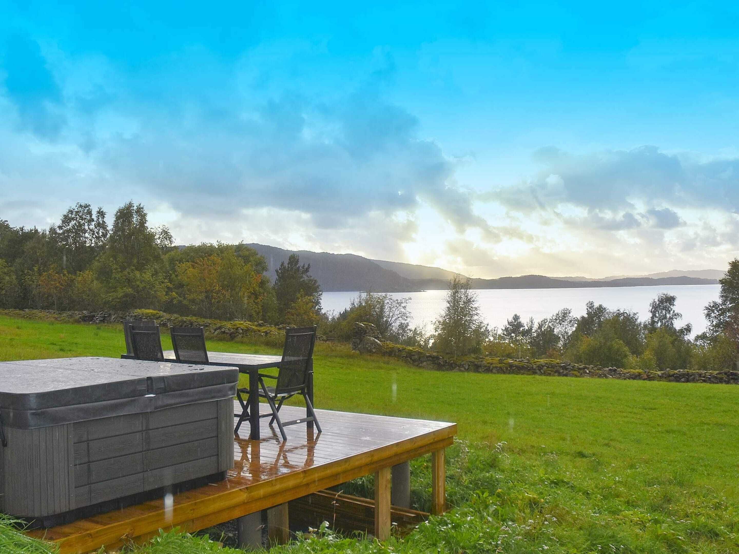 A hot tub on a wooden deck with chairs, overlooking a lake and hills.