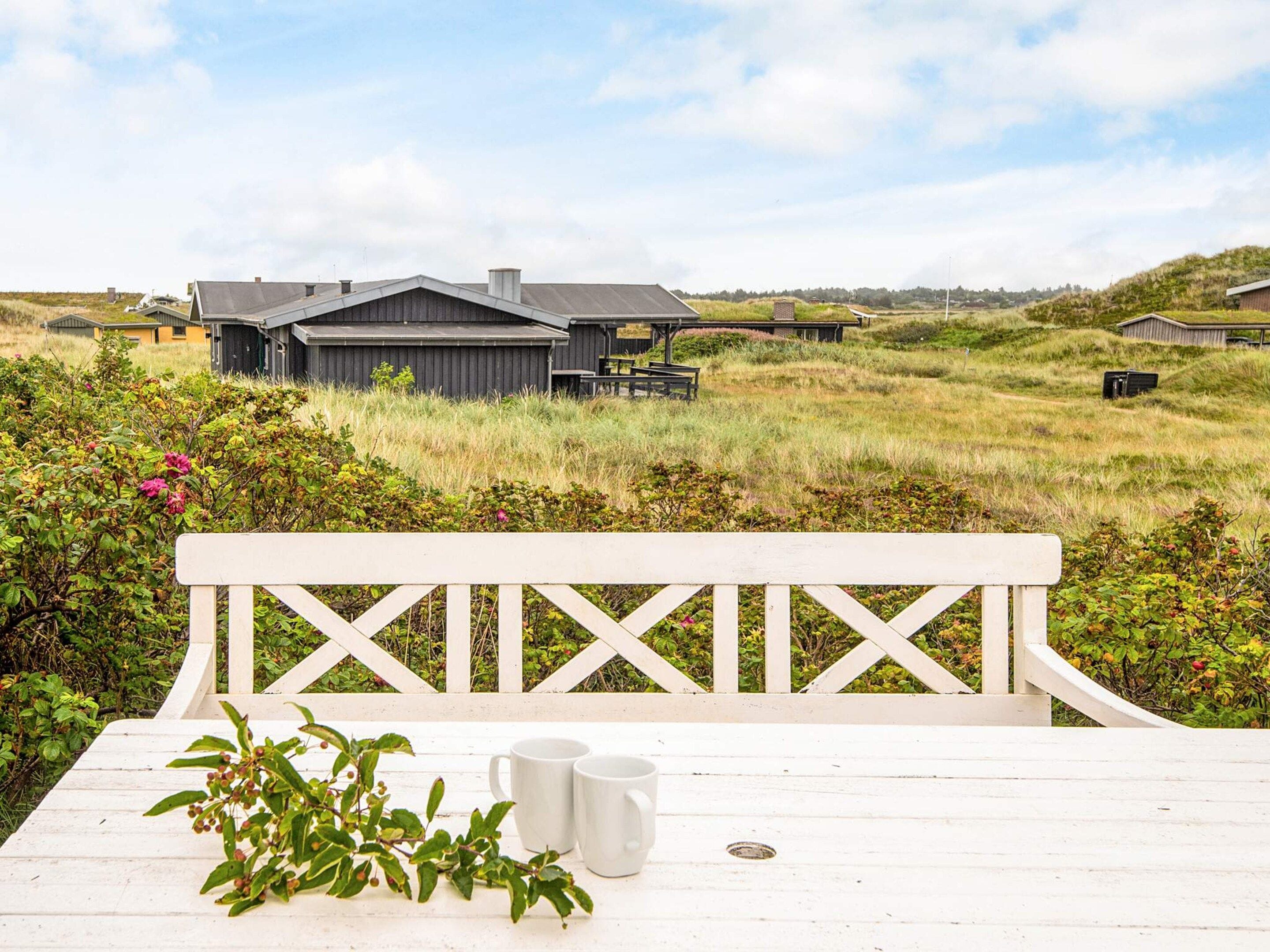 Een witte houten tafel met twee witte mokken en een bloempot, voor een achtergrond van een grasveld en een rij huizen.