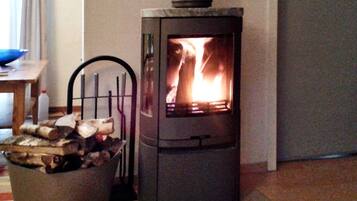 A wood-burning stove with visible flames, a chair, a table with a bottle, and a basket of logs.