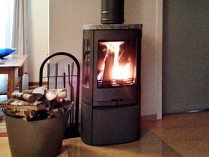 A wood-burning stove with visible flames, a chair, a table with a bottle, and a basket of logs.