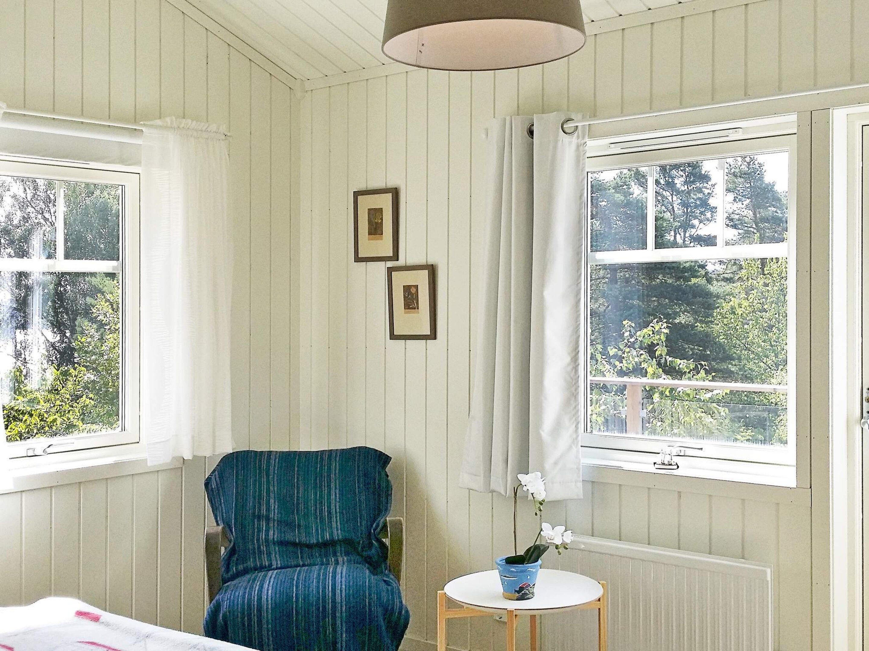 A room with two windows, a chair, a small table with a potted plant, and two framed pictures on the wall.