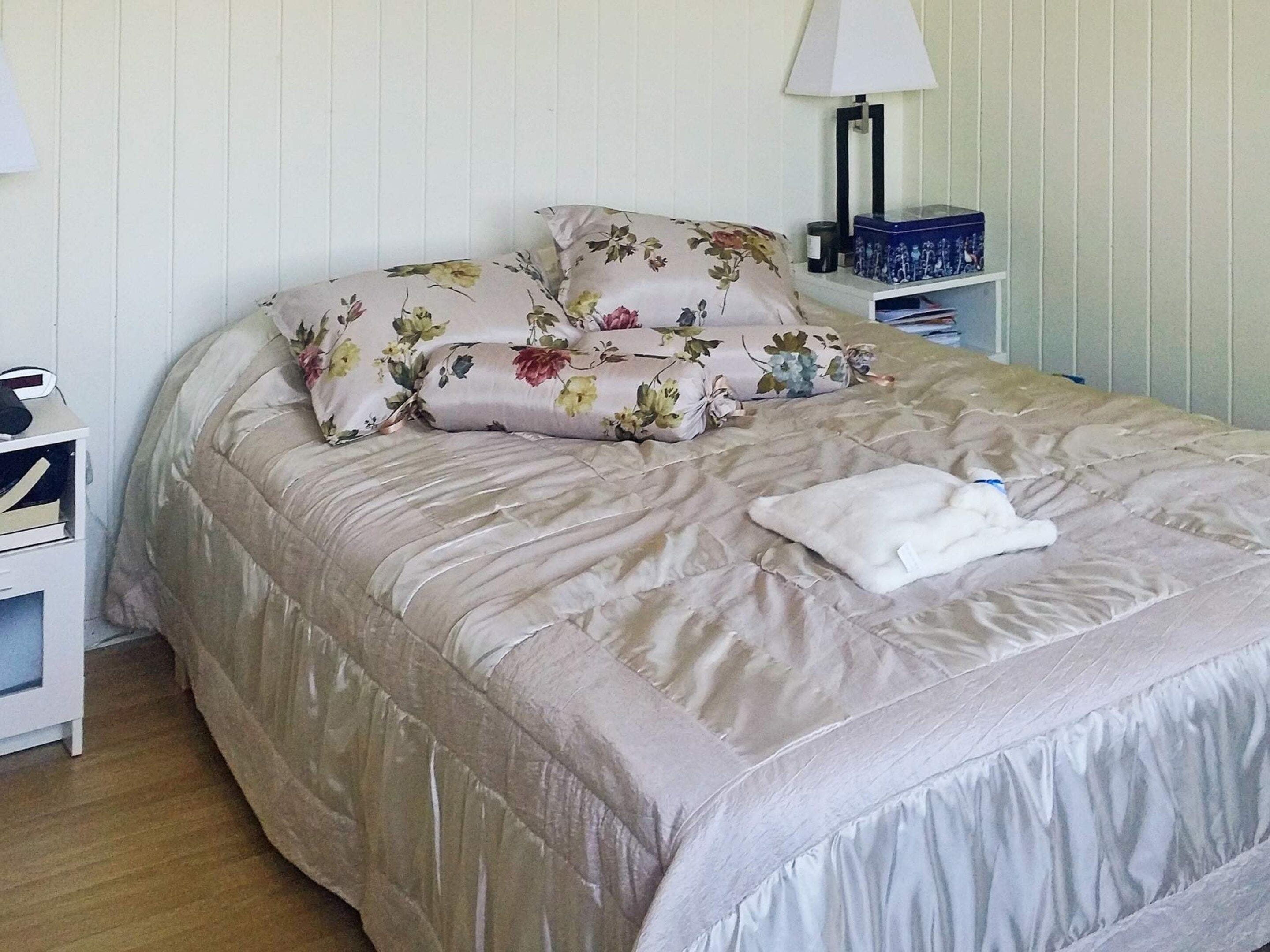 Een netjes opgemaakt bed met bloemenbedrukking, een bedlampje en een klein nachtkastje met een lamp en een doos.