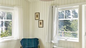 Una habitación con dos ventanas, una silla, una mesita con planta en maceta y dos cuadros en la pared.
