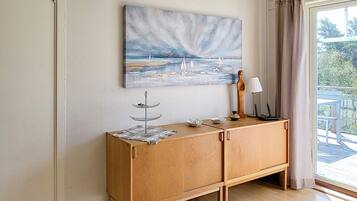 A room with a wooden cabinet, a painting of a seascape, a lamp, and a view of the outdoors through a window.