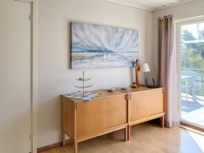 A room with a wooden cabinet, a painting of a seascape, a lamp, and a view of the outdoors through a window. - 4 Person Holiday Home in Uddevalla (Uddevalla)