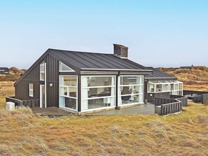 Een modern huis met een grote glazen gevel, een houten terras en een metalen dak.