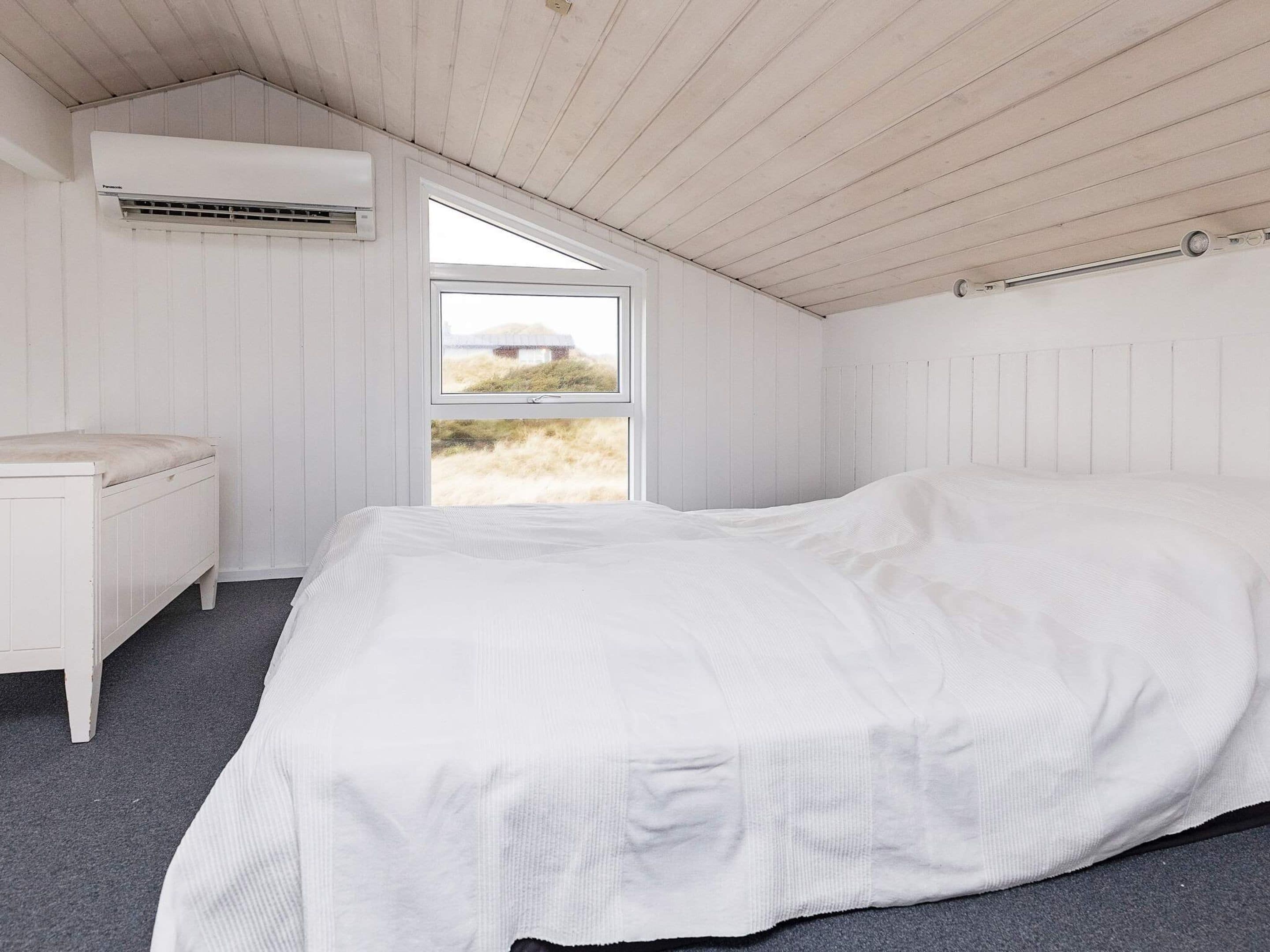 A bedroom with a bed, a nightstand, an air conditioning unit, and a window with a view of the outdoors.