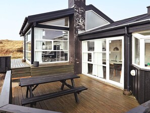 Une terrasse en bois avec une table de pique-nique, un bâtiment à plusieurs fenêtres dont la structure est en verre, et un mur extérieur de couleur noire.