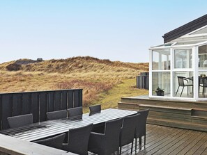 A wooden deck with a table and chairs, a glass-enclosed room with a view of the dunes, and a vehicle in the background.
