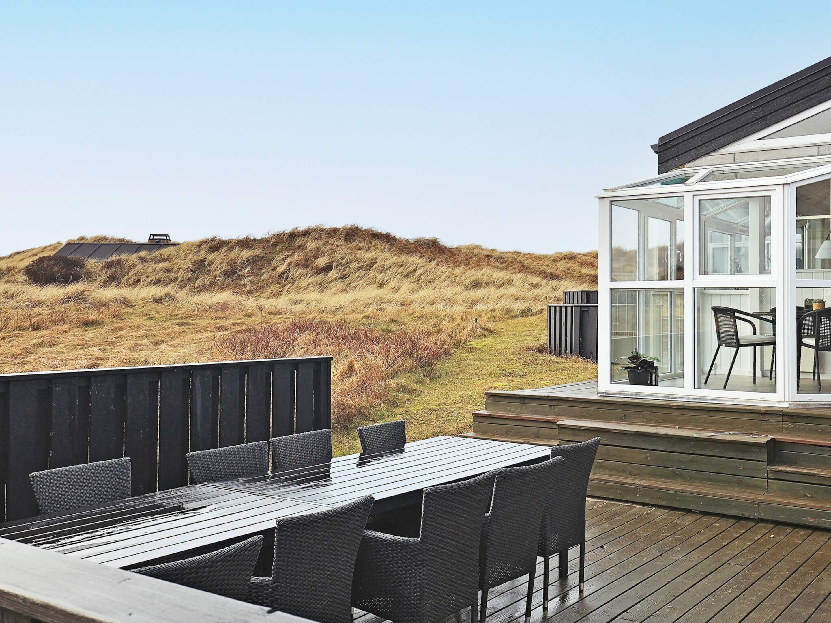 Een houten terras met een tafel en stoelen, een kamer met glazen wanden en uitzicht op het landschap, en een heuvel op de achtergrond.