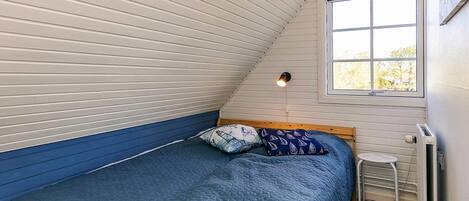 A bedroom with a sloped ceiling, a bed with a blue quilt, a small stool, and a radiator.