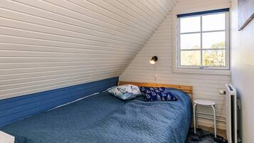 Ein Schlafzimmer mit geneigter Decke, einem Bett mit blauer Bettdecke, einem kleinen Hocker und einem Fenster mit Blick auf Bäume.