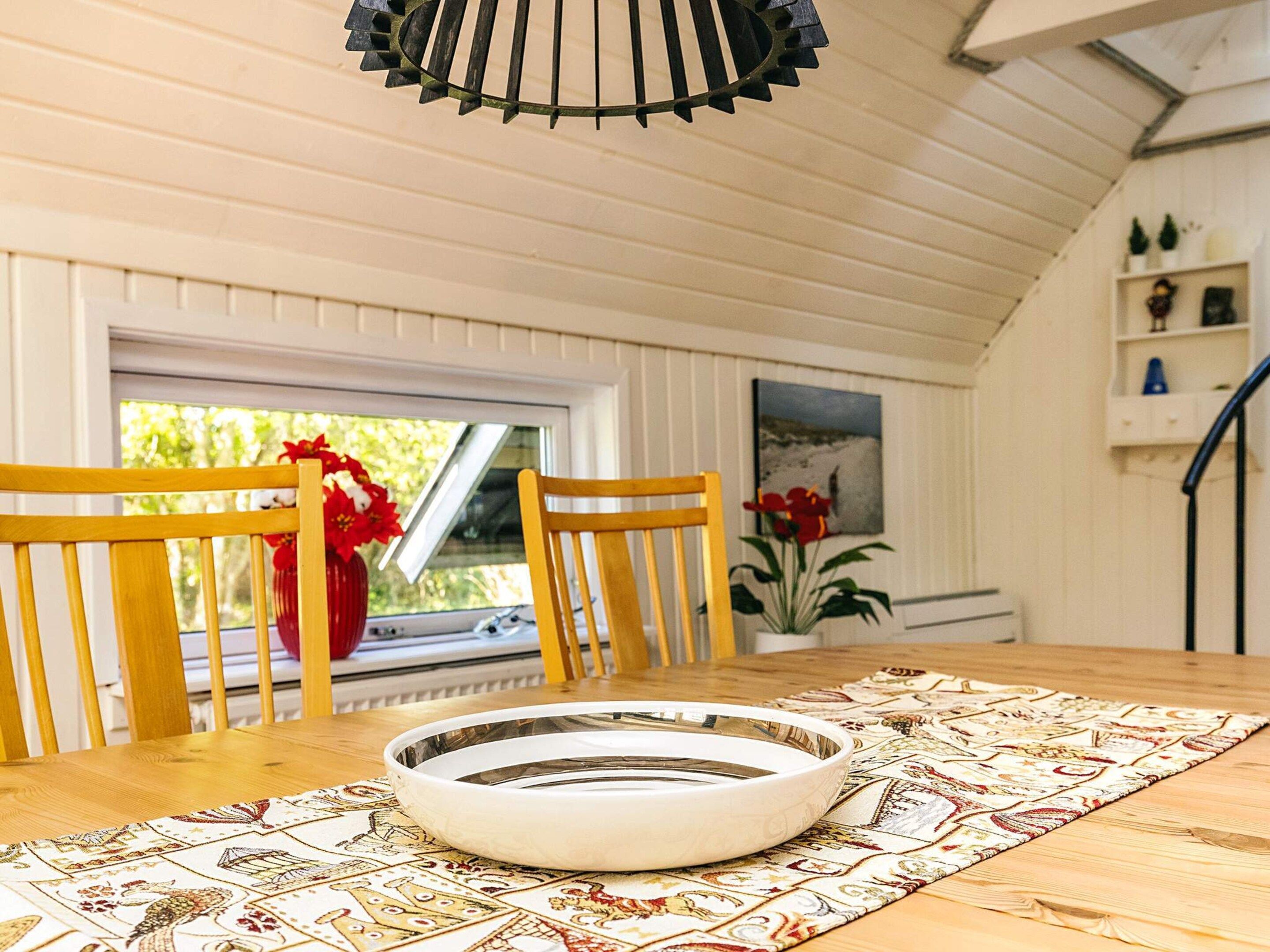 A dining area with a wooden table, two chairs, a decorative plate, a patterned table runner, a window with a view of greenery, and a ceiling fan.