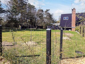 A fenced yard with a grassy area, trees, and a building with a dark roof and brick chimney.