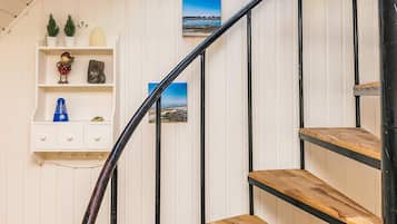 Un escalier avec des marches en bois et des rampes en métal noir, menant à un palier avec un mur blanc et une étagère ornée d’objets décoratifs.