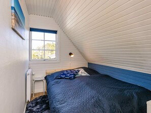 Une petite pièce accueillante avec un plafond en pente, un lit simple recouvert d’une courtepointe bleue, une petite fenêtre donnant sur un paysage agréable et un luminaire fixé au mur.