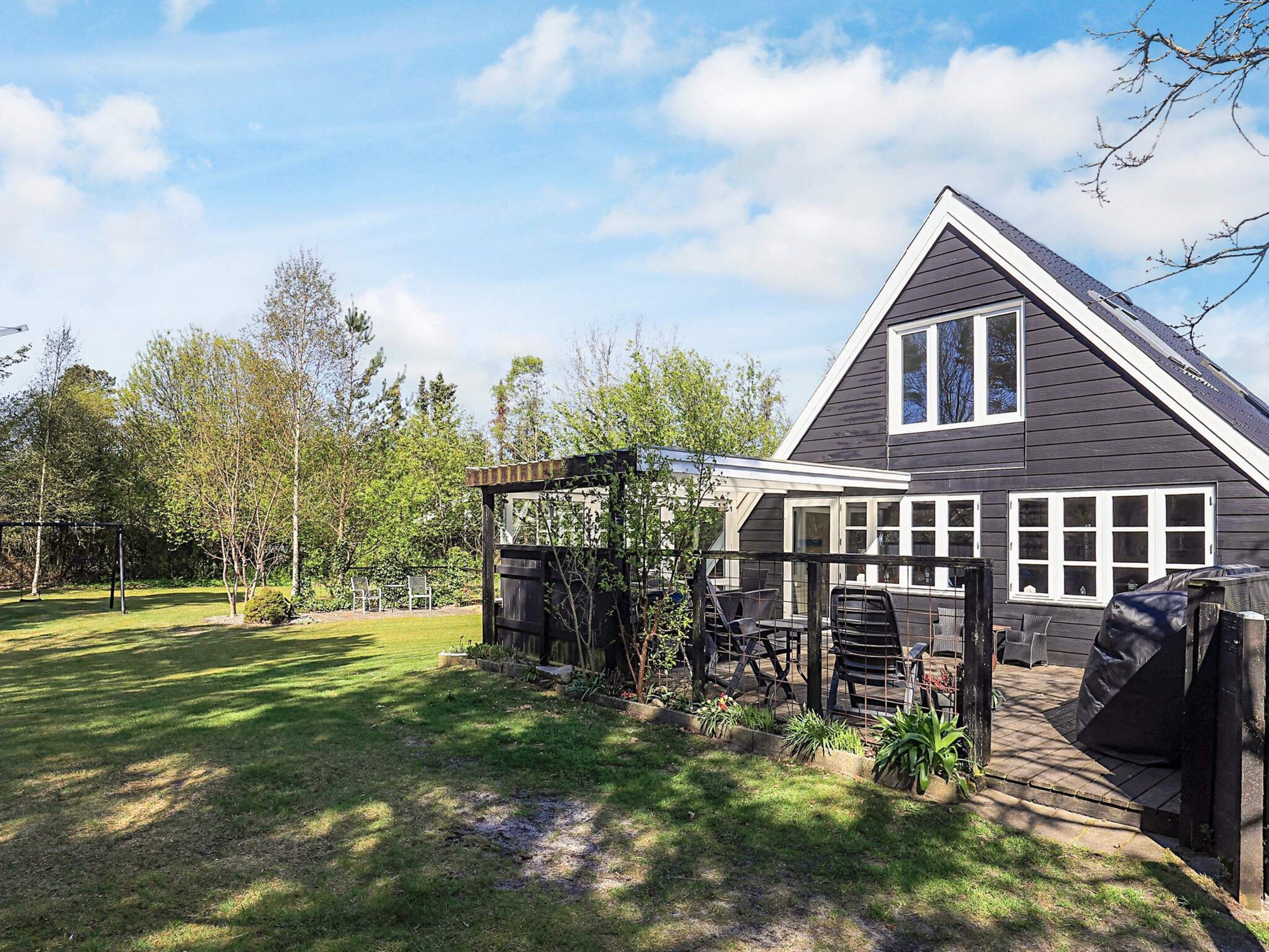 Ein Haus mit dunkelgrauer Fassade, weißem Zierdekor und einem großen Fenster. Eine Terrasse mit Gartenmöbeln und einer Pergola ist ebenfalls vorhanden.