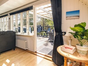 A cozy room with a large window, a radiator, a grey sofa, a wooden table with a box and a plant, and a view of an outdoor deck with chairs and a table. - 6 Person Holiday Home in Hals-by Traum (Hals)