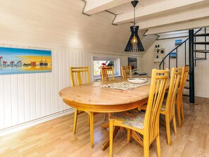 A dining area with a round wooden table, six wooden chairs, a black hanging light fixture, and a painting of a coastal scene on the wall.
