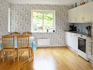 Une cuisine avec des meubles blancs, une fenêtre donnant sur un espace verdoyant et un coin repas avec des chaises et une table en bois.