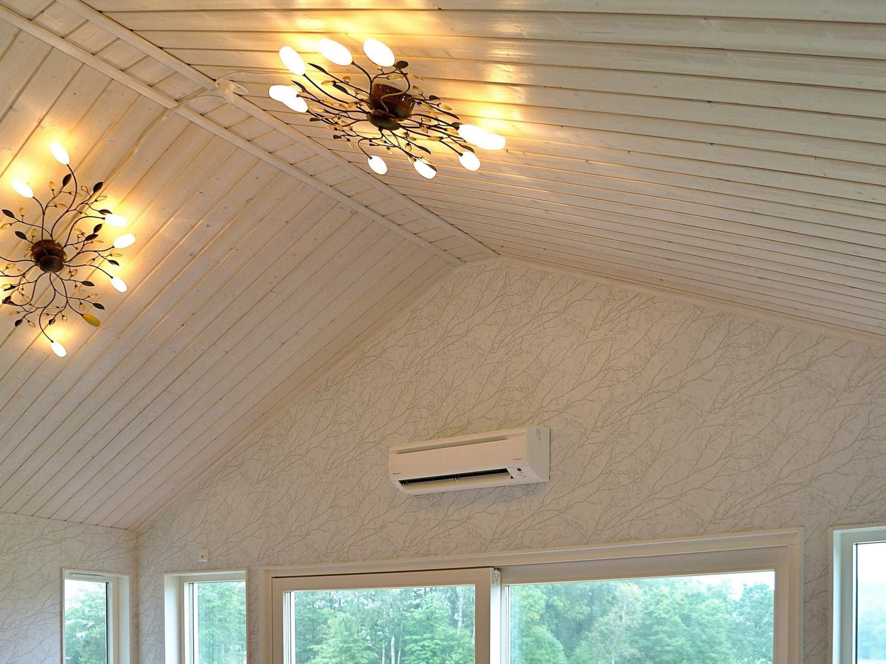 A room with a wooden ceiling, a decorative light fixture, an air conditioning unit, and large windows with a view of trees.