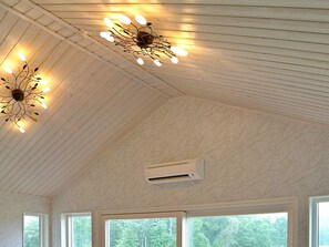 Une pièce avec un plafond en bois, un luminaire décoratif, un appareil de climatisation et de grandes fenêtres donnant sur la forêt.