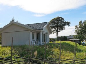 A single-story building with a gabled roof, a porch with two chairs, and a large tree in the background.