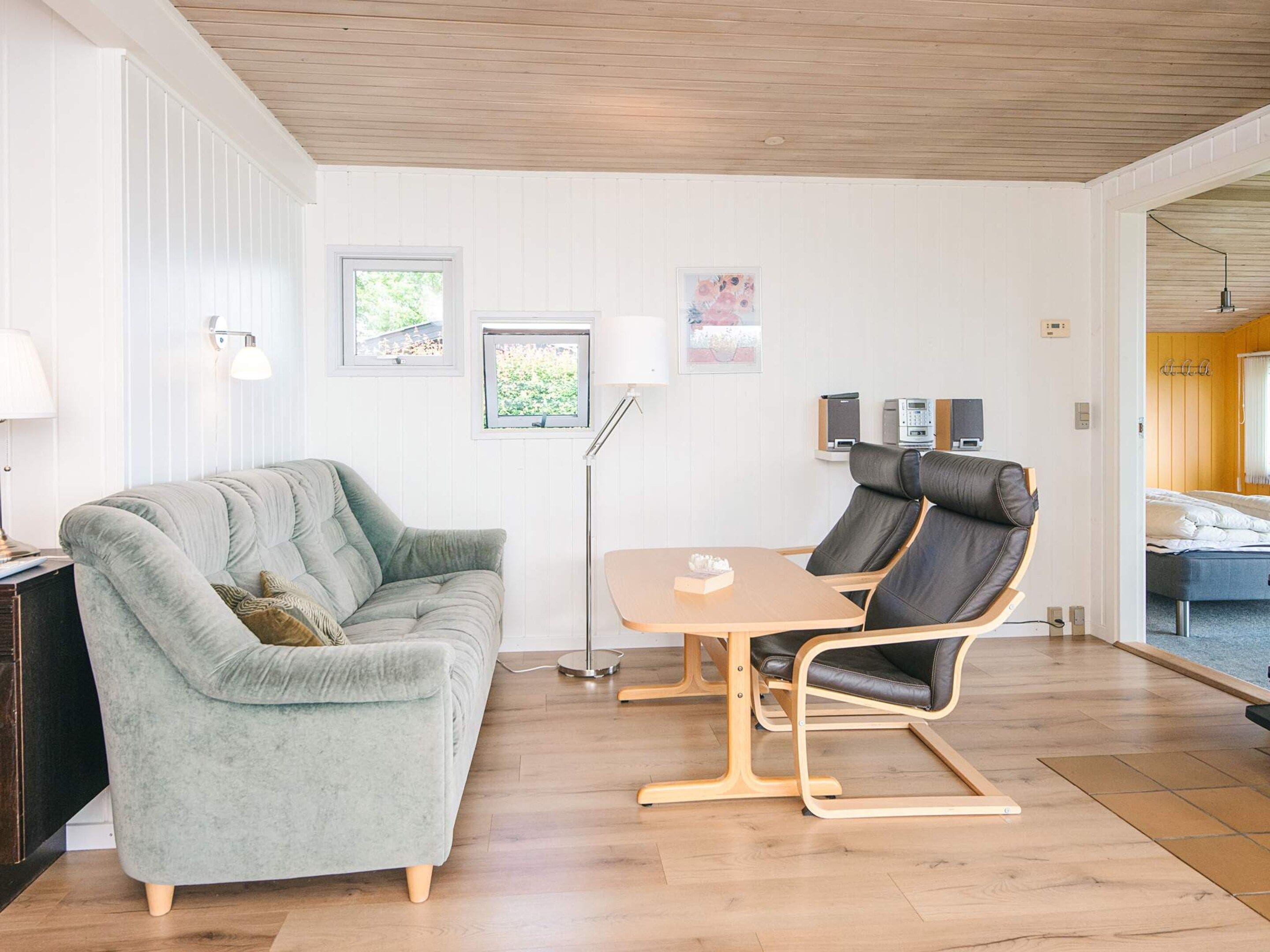 A modern living room with a grey sofa, a wooden coffee table, and a black leather chair. There are two windows with curtains, a floor lamp, and a small table with a book.