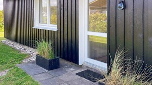 Un edificio moderno con fachada oscura, una ventana con marco blanco y una puerta con panel de vidrio. Hay dos macetas negras con césped en el suelo.