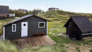 A small, dark wooden cabin with a white door and windows, a wooden deck, and a grassy landscape with hills in the background. - 5 Person Holiday Home in Ringkobing-by Traum (Ringkobing)
