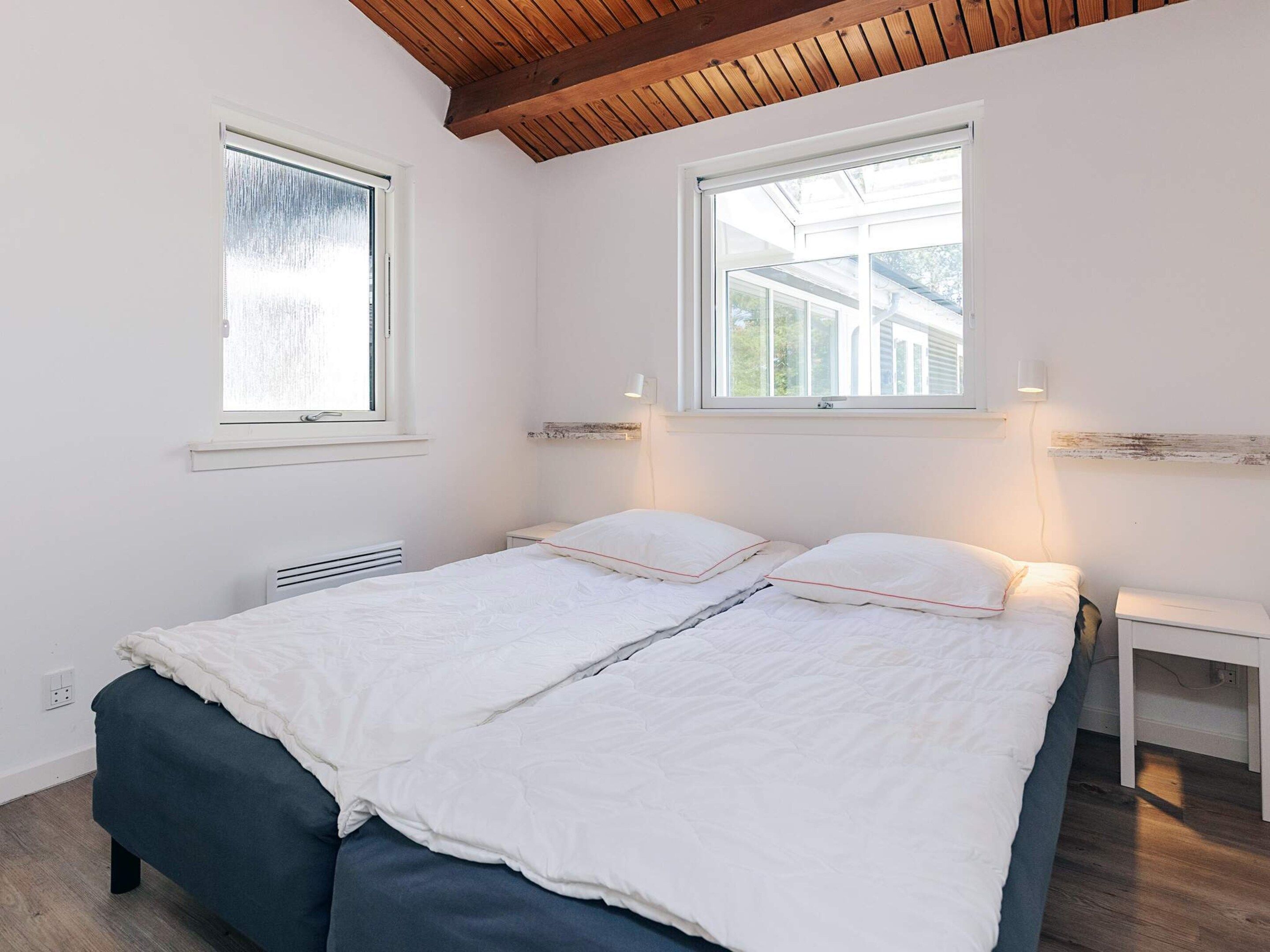 A bedroom with a bed, two windows, a bedside table, and a wooden ceiling.
