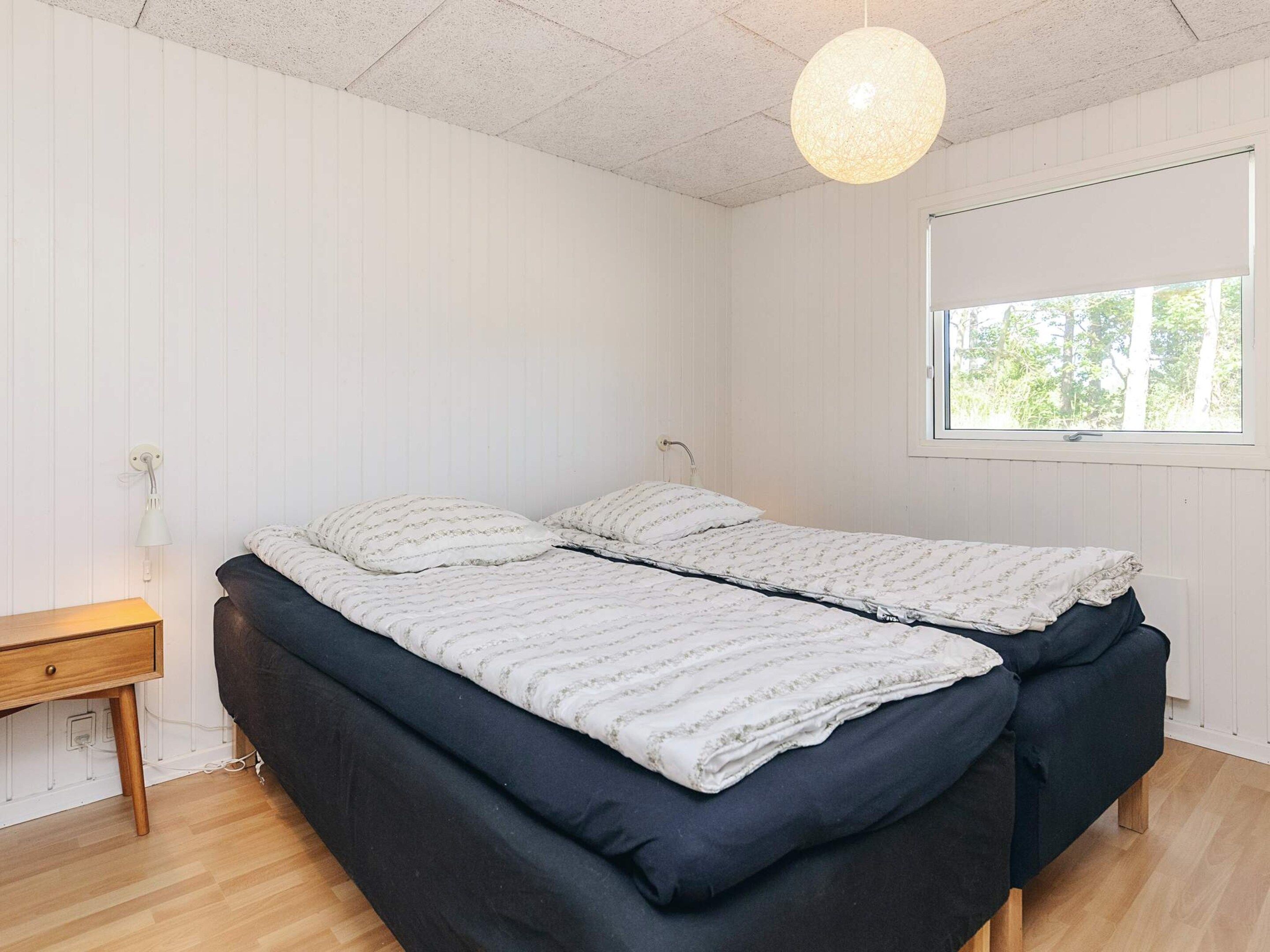 A bedroom with a double bed, a bedside table, a hanging light fixture, and a window with a view of trees.