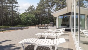 Eine Terrasse mit weißen Kunststoffliegen, einem Grillbereich und einem Gebäude mit Glasfassade und einer Schiebetür.