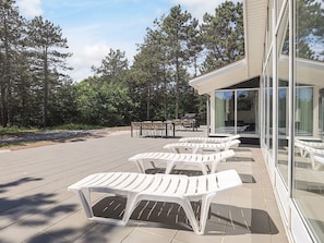 Une terrasse dotée de chaises longues en plastique blanc, d’un espace barbecue et d’un bâtiment clos par des parois de verre, avec une porte coulissante.