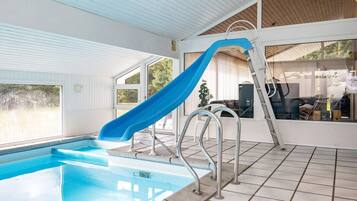 En indendørs swimmingpool med blå rutsjebane, stålrækv og udsigt til det fri gennem store vinduer.
