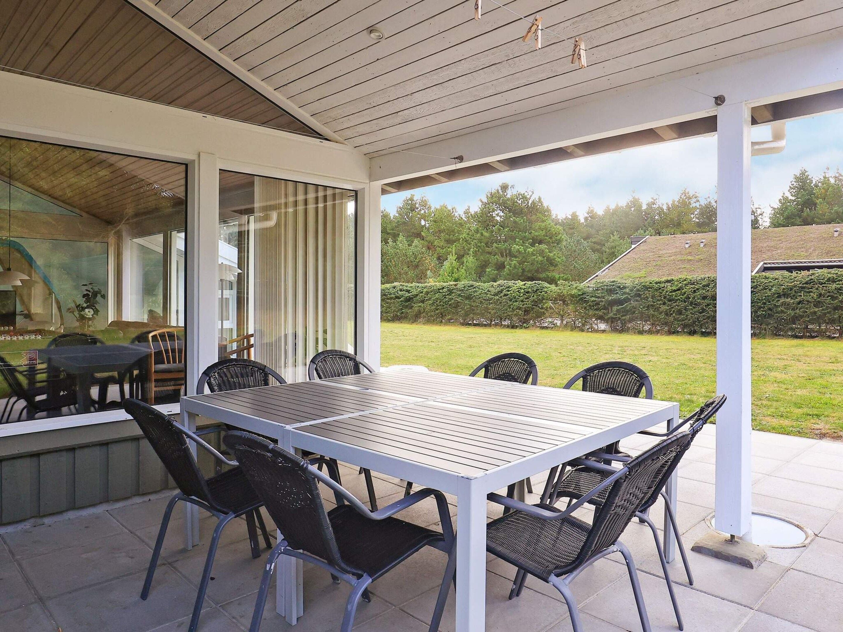 An outdoor patio with a table and chairs, a covered area with a view of a garden and trees.