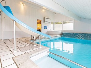 An indoor swimming pool with a blue slide, tiled flooring, and a bench by the pool.