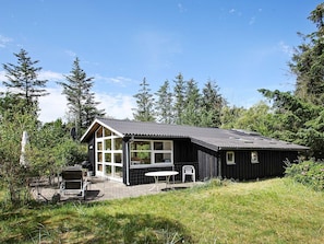 A cabin with a large window, a patio with a table and chairs, surrounded by trees. - Tranquil Holiday Home in Jerup near Sea (Jerup)