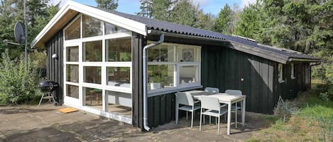 Uma pequena cabana de madeira com uma ampla extensão envidraçada, uma mesa de pátio e uma churrasqueira.