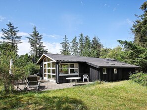 Une cabane dotée d’une grande fenêtre, d’une terrasse avec une table et des chaises, le tout entouré d’arbres.