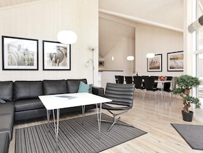 A modern living area with a sofa, a coffee table, and a dining table with chairs. There are framed pictures on the wall, a potted plant, and a lamp.