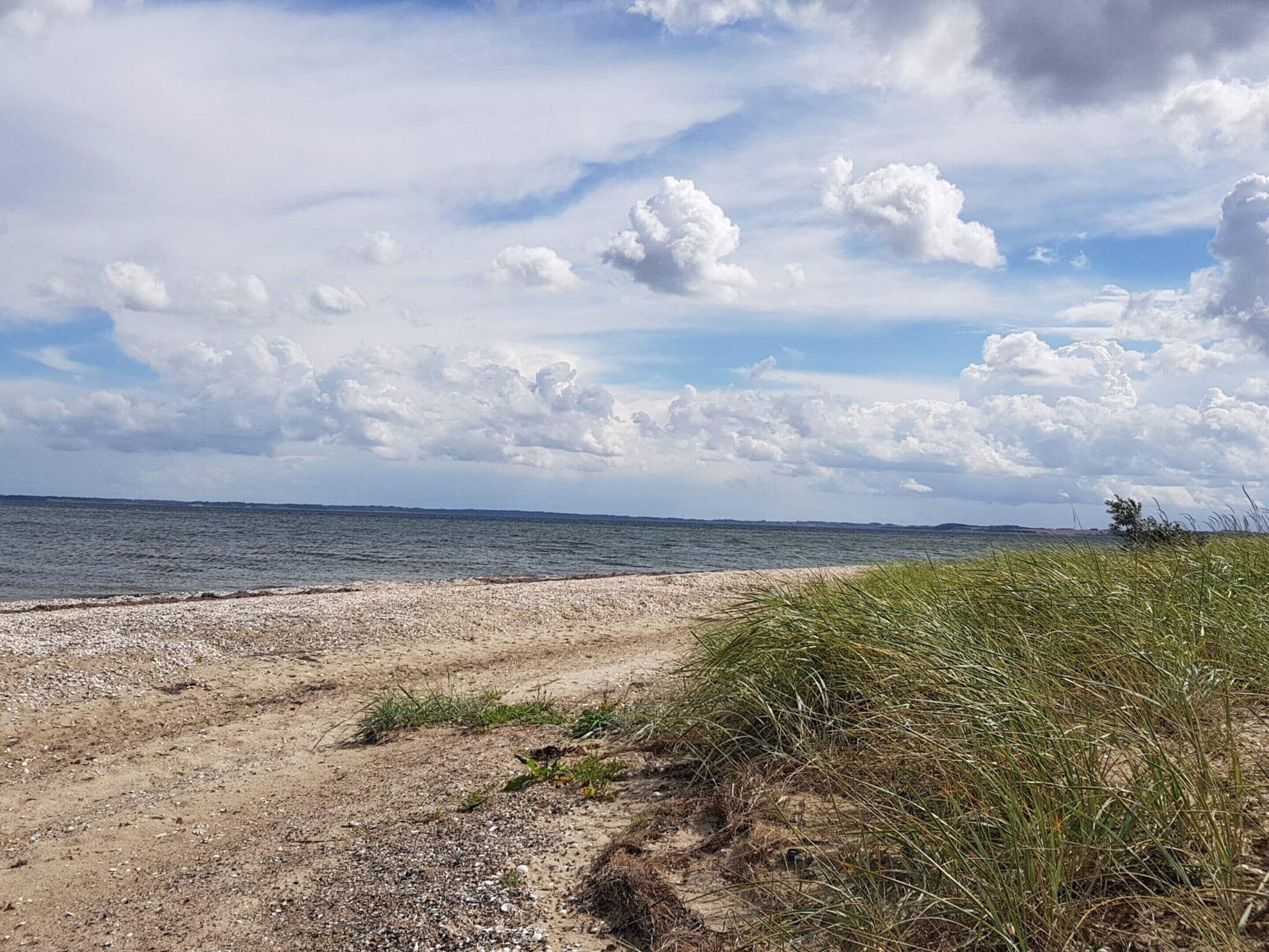 A beach with a pebbly shore, grassy dunes, and a calm sea under a partly cloudy sky.